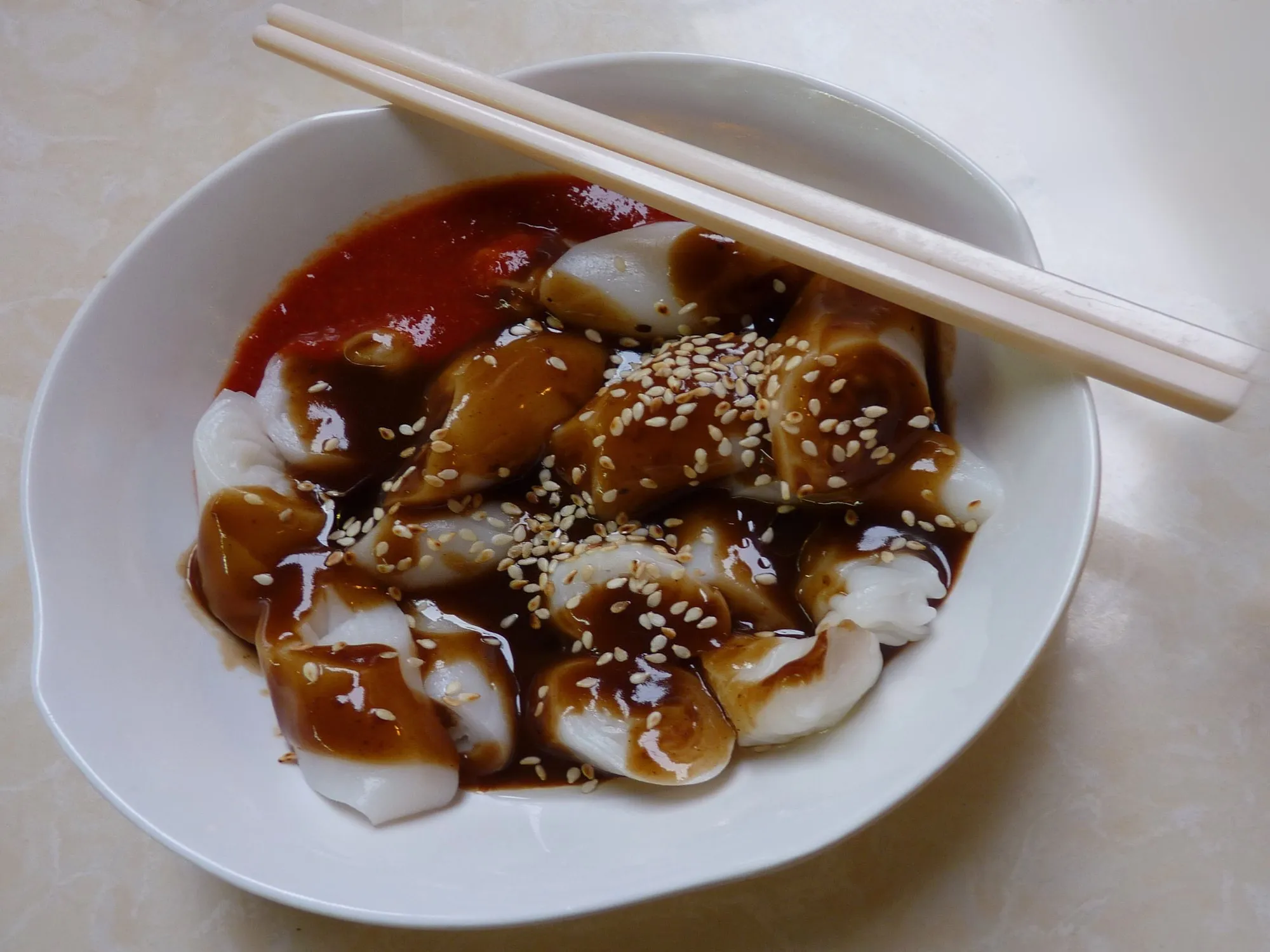Bowl of rolled rice noodle pieces with sweet dark sauce, chilli sauce and sesame seeds