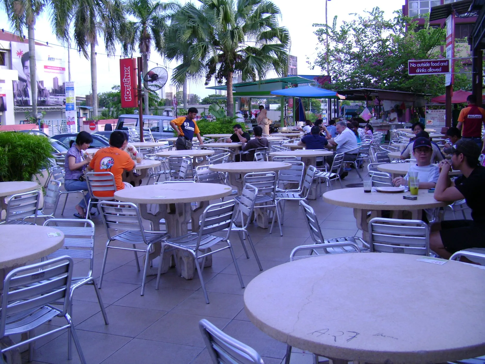 Malaysian coffee-shop seating with rows of round tables, metal chairs and diners under trees