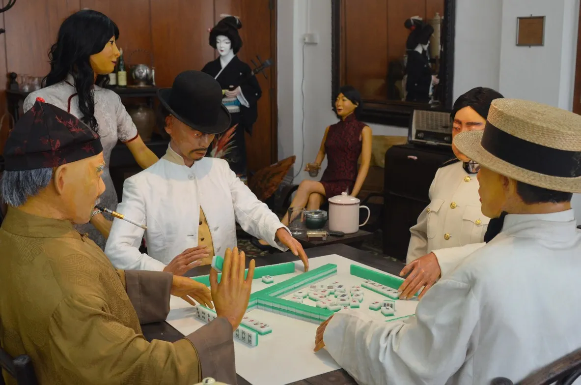 Diorama muzium lelaki berpakaian zaman dahulu bermain mahjong di meja, dengan wanita berdiri di belakang mereka