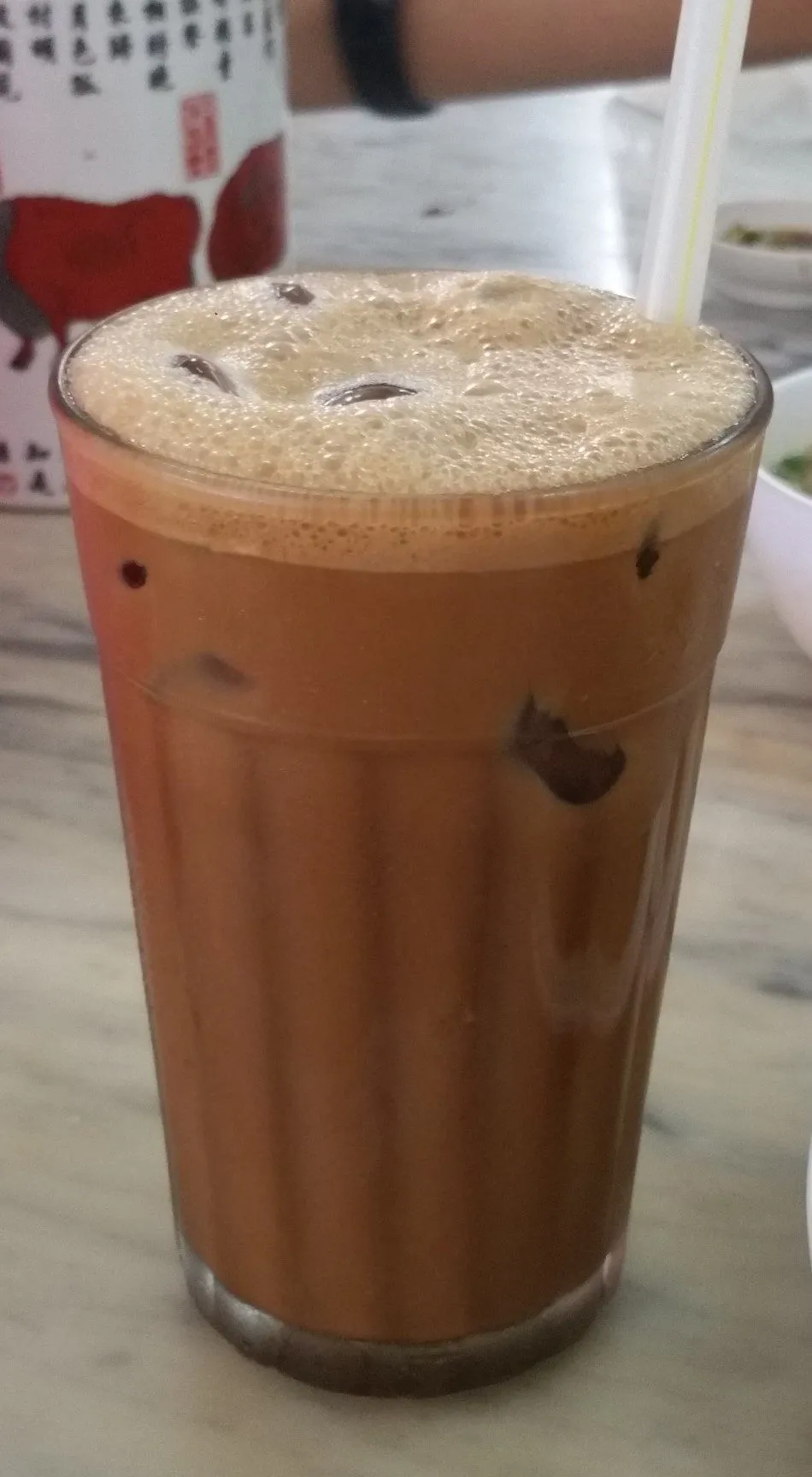 Segelas tinggi kopi putih Ipoh ais berbuih dengan ais dan straw di atas meja marmar