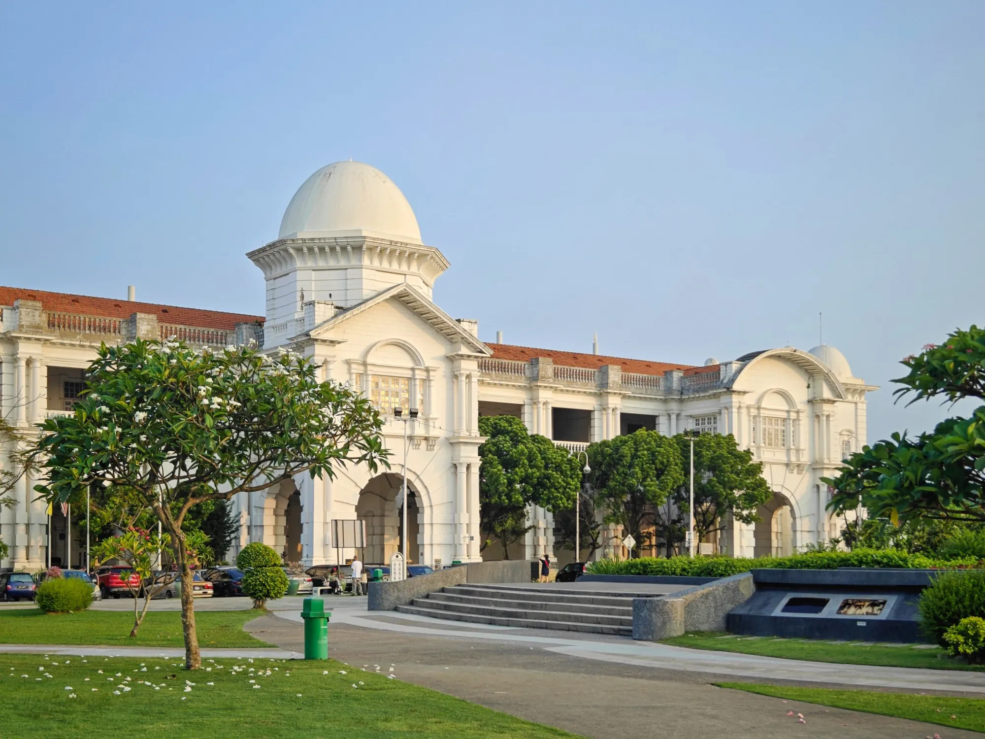 Stesen Keretapi Ipoh era kolonial berwarna putih dengan kubah dan gerbang di belakang halaman rumput dan pokok palma
