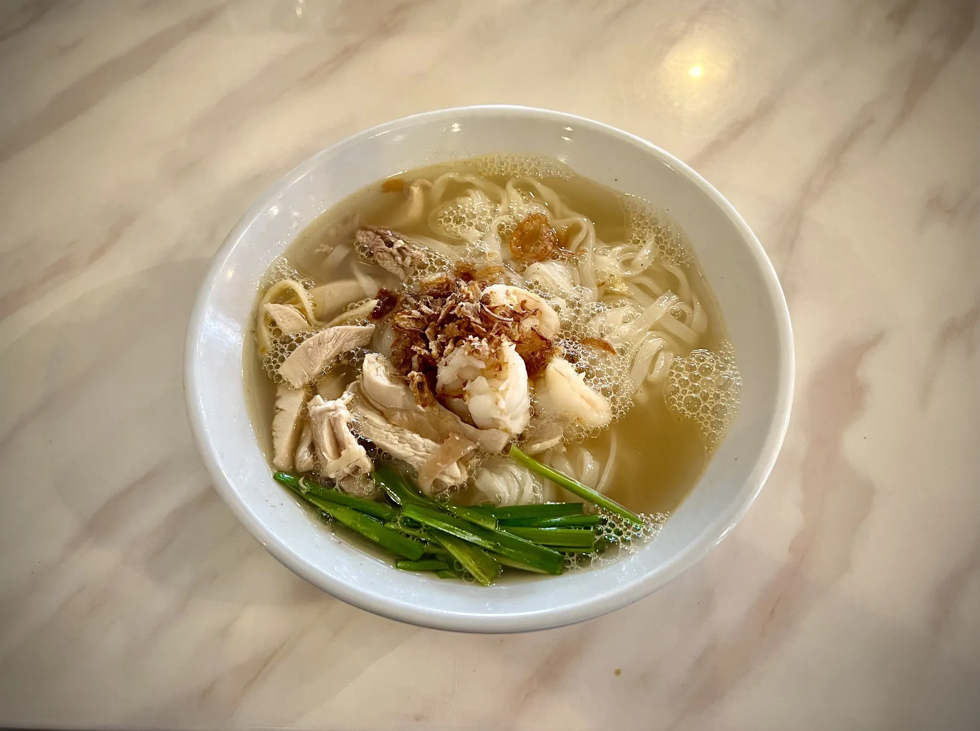 Semangkuk kai si hor fun dengan ayam carik dan kucai dalam sup jernih di atas meja marmar